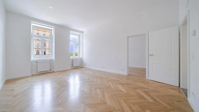 Helles Zimmer mit Fischgrätparkett, weißen Wänden und zwei großen Fenstern.