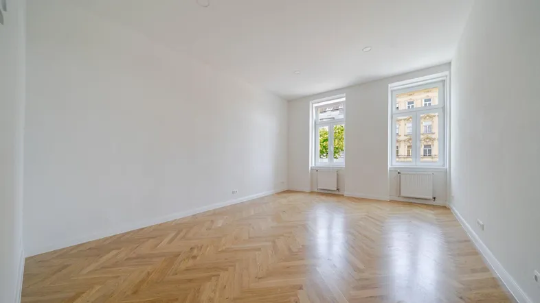 Geräumiges Zimmer mit glänzendem Fischgrätparkett und viel Tageslicht durch zwei Fenster.