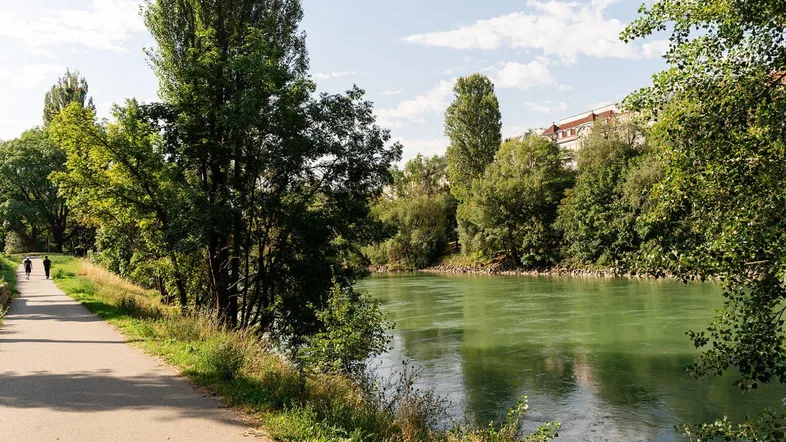 Ein Spazierweg entlang eines grünen Flusses, umgeben von Bäumen und mit Gebäuden im Hintergrund.