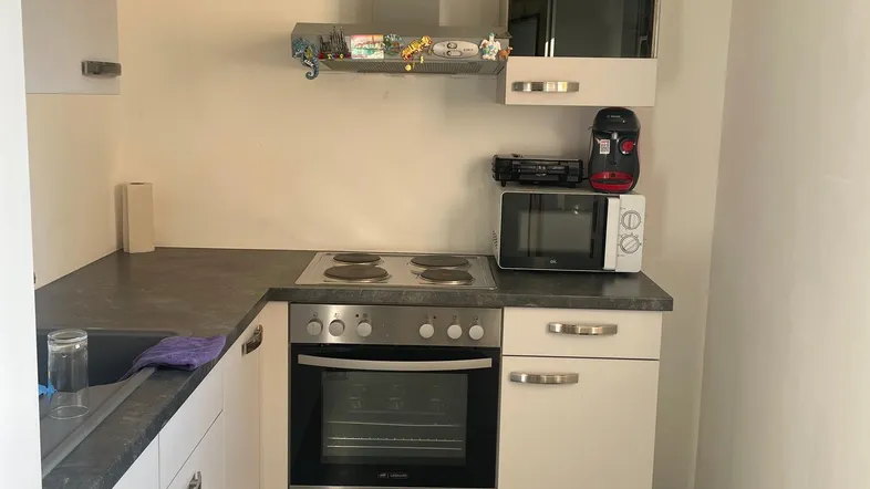 Kleine Einbauküche mit weißen Schränken, dunkler Arbeitsplatte und integriertem Herd mit Backofen.