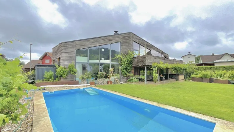 Modernes Haus mit Holzfassade, großem Pool und gepflegtem Rasenbereich unter bewölktem Himmel.