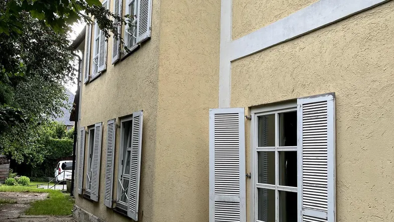 Gelbe Fassade eines älteren Gebäudes mit weißen Fensterläden und einem verwilderten Gartenbereich.