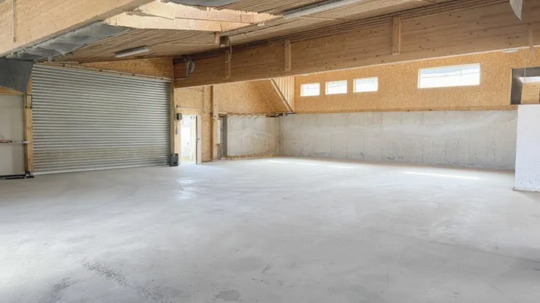 Großer, leerer Lagerraum mit Betonboden, Holzdeckenbalken und einem Rolltor für den Zugang.