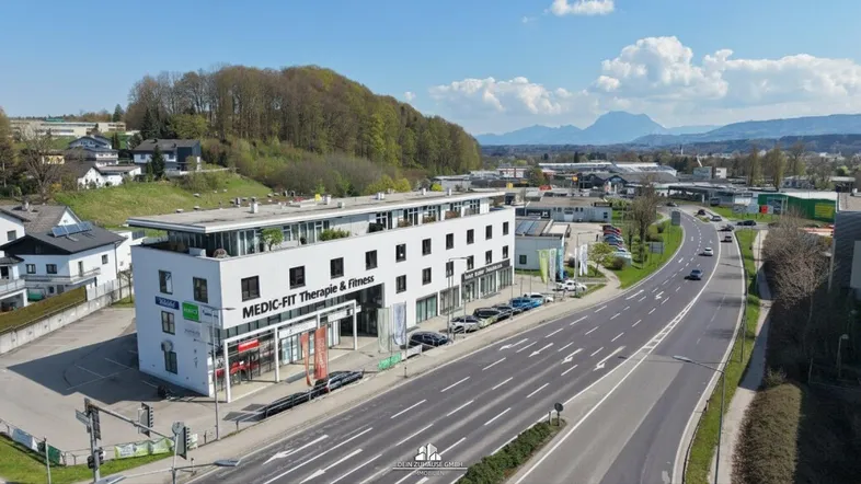 Luftaufnahme eines modernen Geschäftsgebäudes an einer Hauptstraße mit umliegender Landschaft und Bergen im Hintergrund.