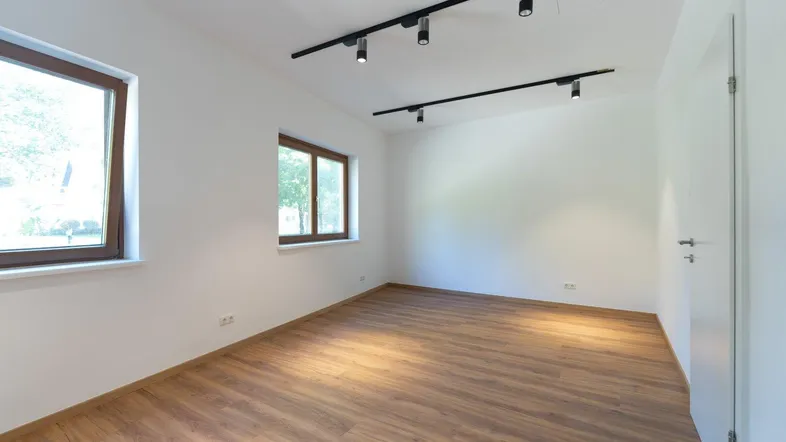 Heller, leerer Raum mit modernem Laminatboden, weißen Wänden und zwei Fenstern mit Blick ins Grüne.