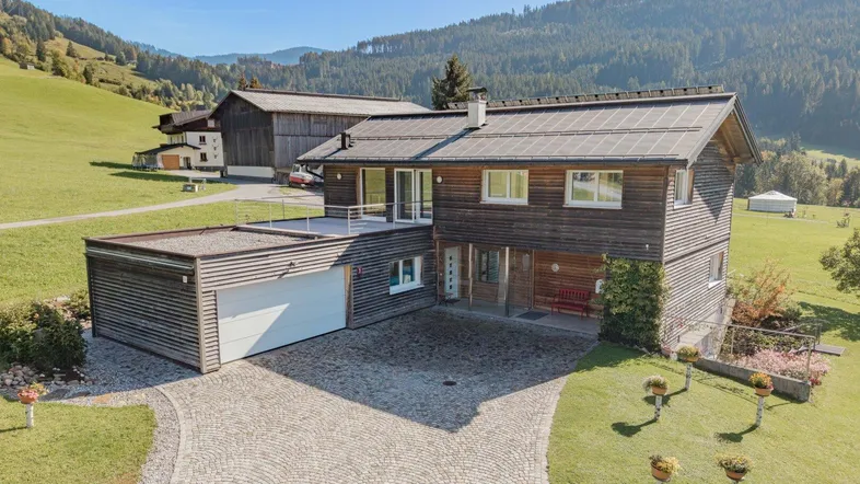 Traditionelles Holzhaus mit integrierter Garage und gepflasterter Auffahrt in alpiner Landschaft.