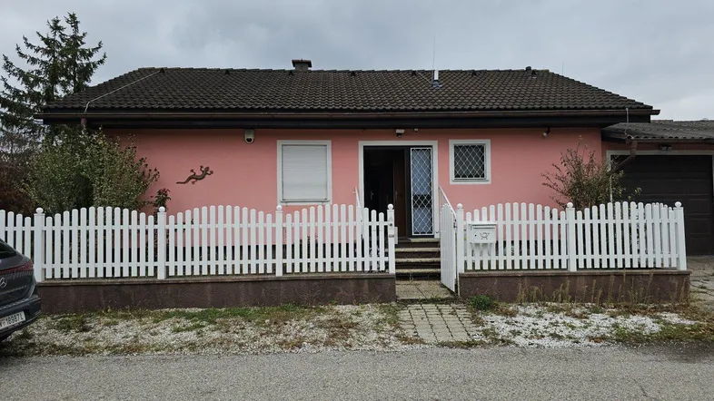 Einfamilienhaus mit rosafarbener Fassade, weißem Lattenzaun und Vorgarten unter bewölktem Himmel.