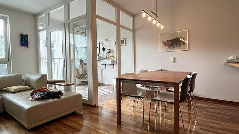 Heller Wohn- und Essbereich mit Holzboden, beigem Sofa und Esstisch, der zum Balkon führt.