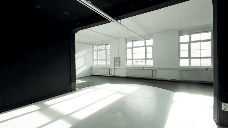 Leerer Innenraum mit großen Fenstern, die viel natürliches Licht hereinlassen und Schattenmuster auf dem Boden erzeugen.