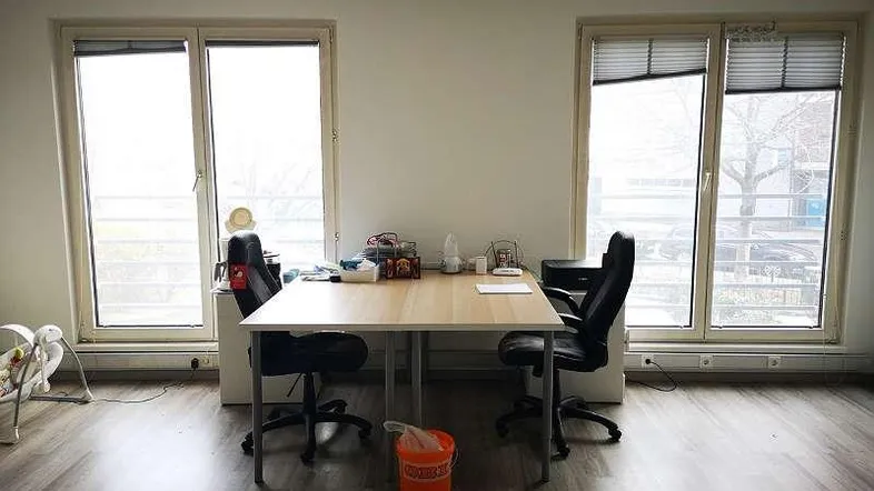 Heller Büroraum mit zwei großen Fenstern, einem Schreibtisch und Bürostühlen.