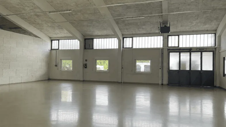 Geräumige, leere Lagerhalle mit hohen Decken, großen Fenstern und einem glänzenden Betonboden.