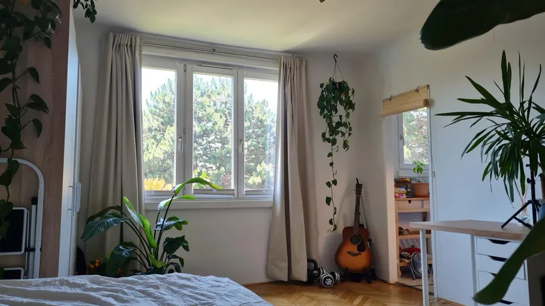 Helles Schlafzimmer mit Parkettboden, vielen Pflanzen und Blick ins Grüne durch das Fenster.