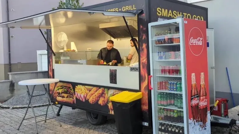 Mobiler Imbisswagen mit offener Theke und Getränkekühlschrank auf gepflastertem Vorplatz.