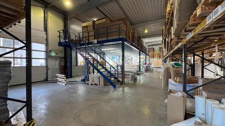 Geräumige Lagerhalle mit hohen Decken, Betonboden und einer Zwischenebene für zusätzliche Lagerfläche und Regale.