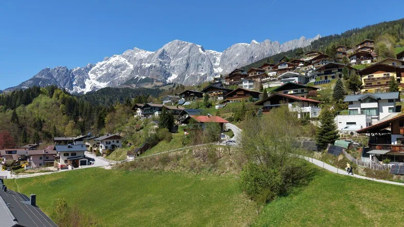 Panoramablick auf ein malerisches Bergdorf mit schneebedeckten Gipfeln im Hintergrund unter blauem Himmel.
