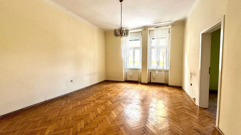 Geräumiges Wohnzimmer mit Fischgrätparkett und zwei großen Fenstern, bereit zur Renovierung.