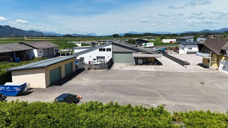 Luftaufnahme eines Gewerbegebiets mit mehreren Gebäuden, großem Parkplatz und Bergkulisse im Hintergrund.