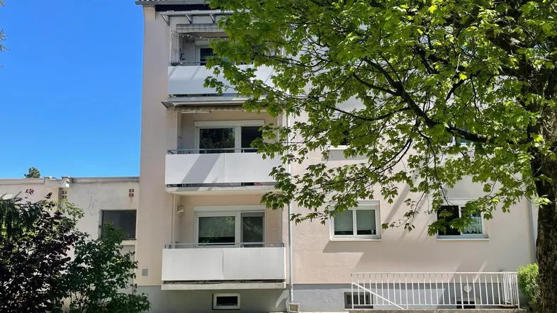 Mehrfamilienhaus mit mehreren Balkonen und grüner Umgebung unter blauem Himmel.