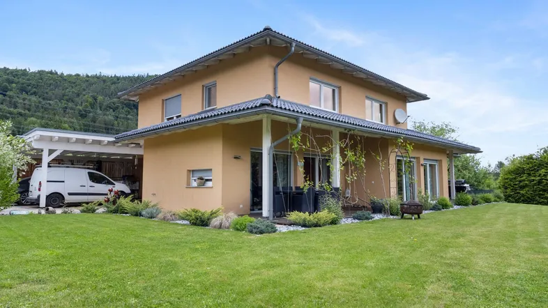 Modernes Einfamilienhaus mit großem Garten, Carport und Blick auf die umliegende Natur.