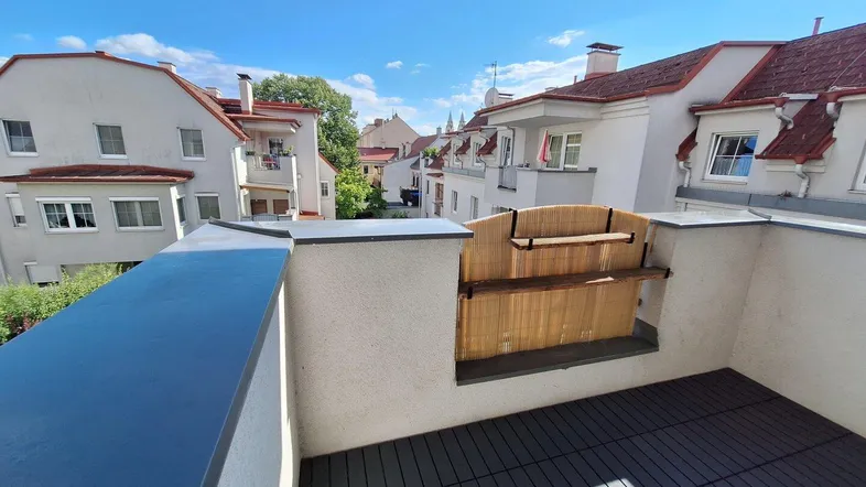 Heller Balkon mit grauen Bodenfliesen und Blick auf die umliegenden Wohngebäude unter blauem Himmel.
