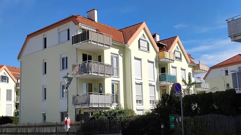 Mehrfamilienhaus mit hellgelber Fassade, roten Ziegeldächern und mehreren Balkonen unter blauem Himmel.