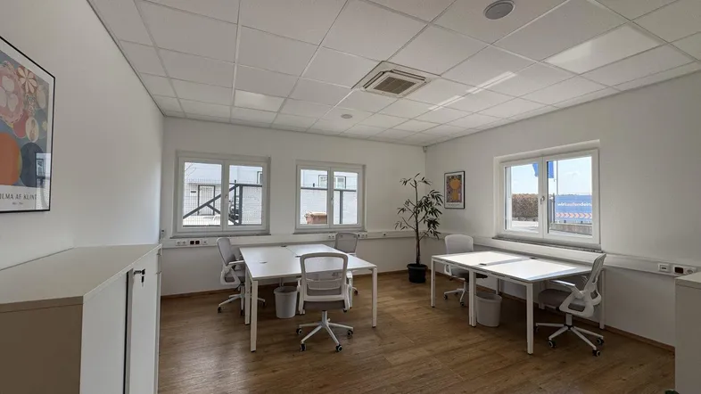 Helles und modernes Büro mit mehreren Arbeitsplätzen, Holzboden und großen Fenstern, die viel Tageslicht hereinlassen.