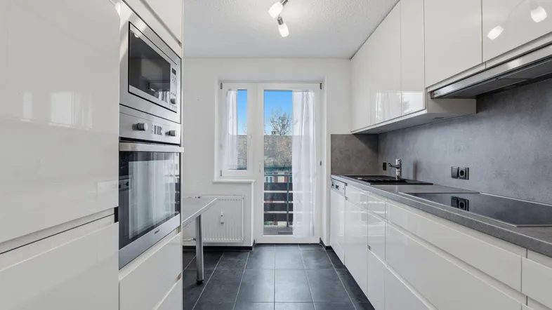 Moderne Küche mit weißen Hochglanzfronten, dunklen Fliesen und Zugang zum Balkon.