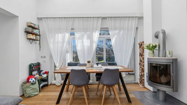 Heller Essbereich mit Holztisch, Kamin und großen Fenstern, die viel Tageslicht hereinlassen.