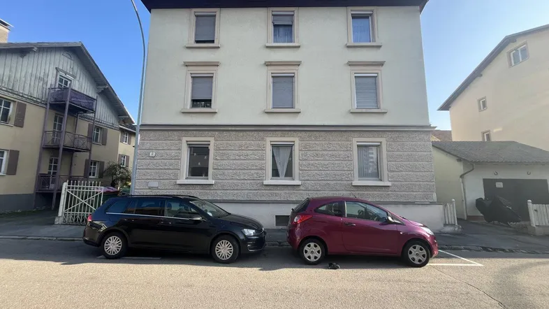 Mehrstöckiges Wohngebäude mit heller Fassade und geparkten Autos auf der Straße davor.