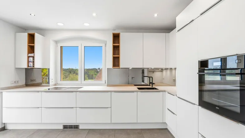 Moderne weiße Küche mit Einbaugeräten, Fliesenboden und einem Fenster mit weitem Blick ins Grüne.