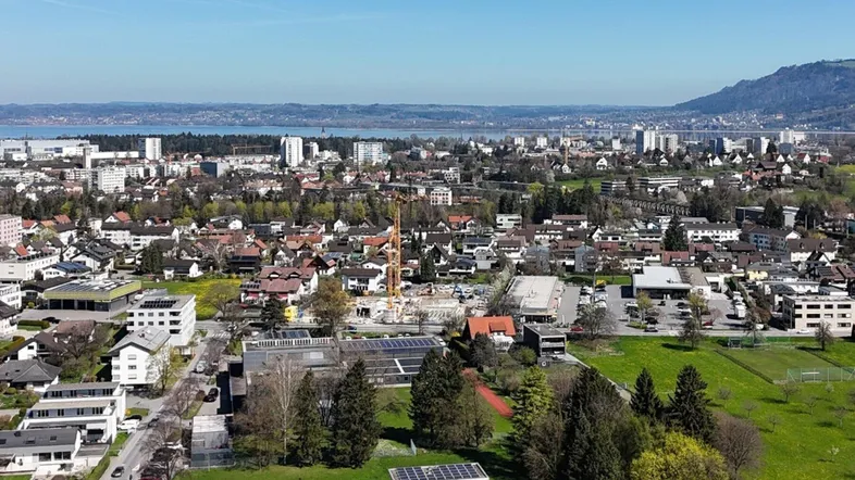 Luftaufnahme einer Stadt mit Wohngebäuden, Grünflächen, einem See und Bergen im Hintergrund.