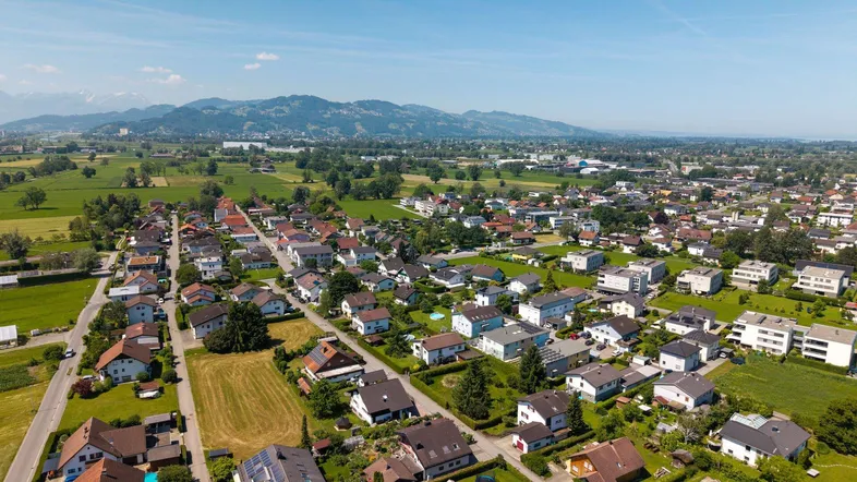 Weitläufige Luftaufnahme einer Wohnsiedlung mit grünen Feldern und Bergen im Hintergrund.