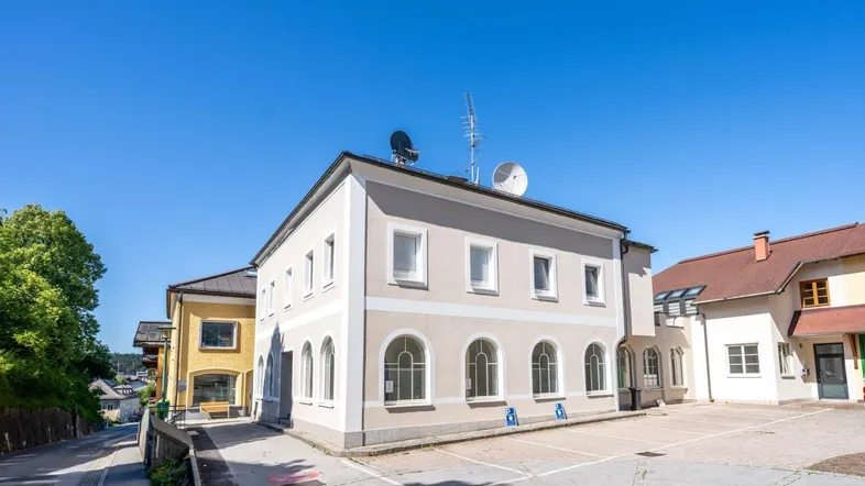 Gebäudeansicht mit heller Fassade, Bogenfenstern und Parkplätzen vor dem Eingangsbereich.