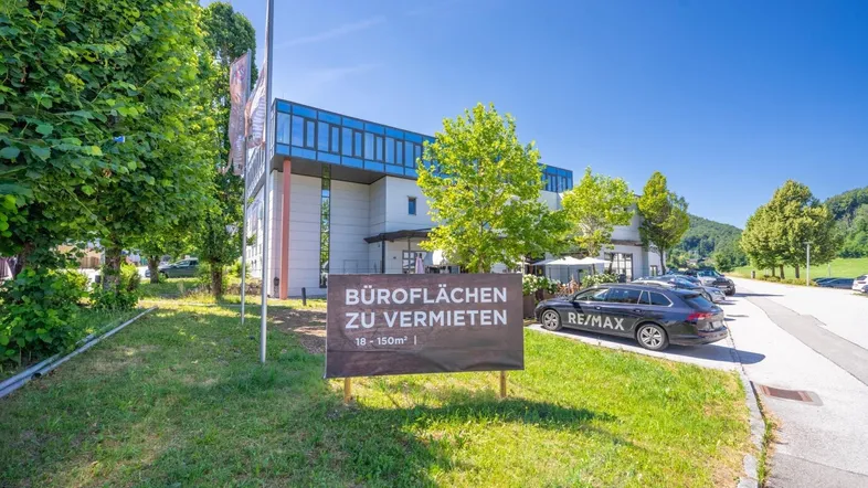 Modernes Bürogebäude mit Glasfassade und Vermietungsschild, umgeben von Grünflächen und Parkplätzen.