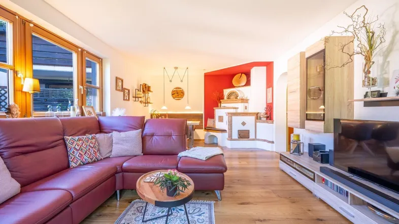 Gemütliches Wohnzimmer mit rotem Ledersofa, Holzboden und traditionellem Kachelofen, hell durch große Fenster.