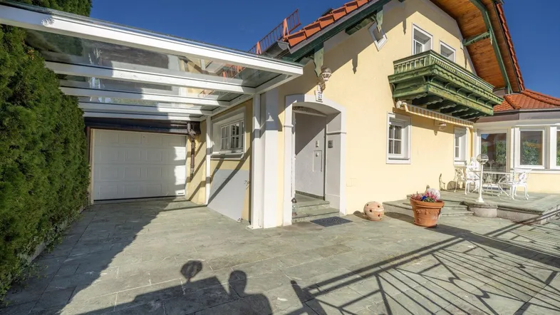 Eingangsbereich des Hauses mit überdachtem Carport und gepflastertem Vorplatz unter blauem Himmel.