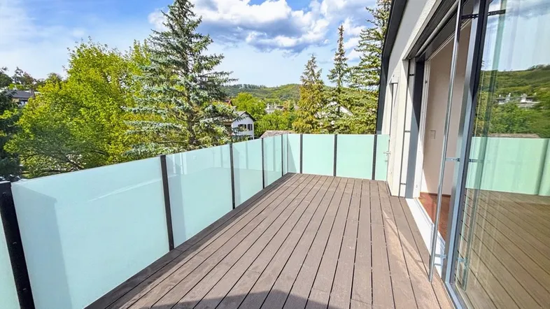 Geräumiger Balkon mit Holzboden und Glasbrüstung, umgeben von grüner Natur und Bäumen.