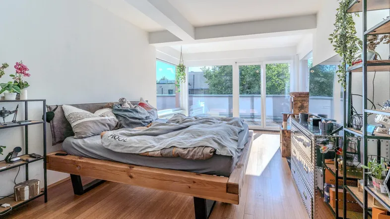 Helles Schlafzimmer mit Holzbett, Parkettboden und großen Fenstern, die viel Sonnenlicht hereinlassen.