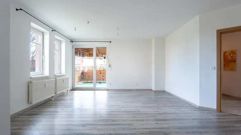 Helles, leeres Wohnzimmer mit Laminatboden und Zugang zu einer überdachten Terrasse.