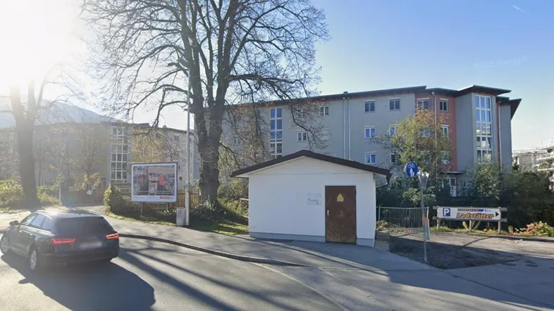 Ansicht eines mehrstöckigen Wohngebäudes mit umliegenden Bäumen, einer Straße und einem kleinen Nebengebäude in sonniger Umgebung.