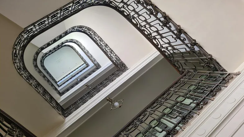 Blick von unten in ein historisches Treppenhaus mit kunstvollen schmiedeeisernen Geländern und hohen Decken.