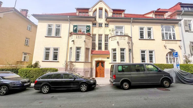 Historisches Gebäude mit beiger Fassade, Erkern und parkenden Autos auf der Straße.