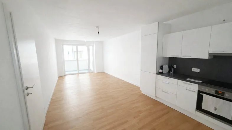 Heller offener Wohnbereich mit integrierter Küche, Holzboden und Zugang zum Balkon.