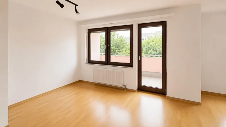 Helles Zimmer mit Parkettboden, weißen Wänden und Zugang zu einem Balkon.