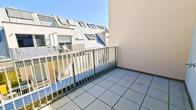 Heller Balkon mit Fliesenboden und Metallgeländer, bietet Blick auf umliegende Gebäude und Dächer.