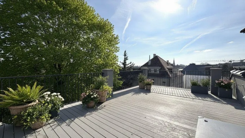 Geräumige Dachterrasse mit Holzdielenboden, Pflanzen und weitem Blick über die Stadt und grüne Bäume.