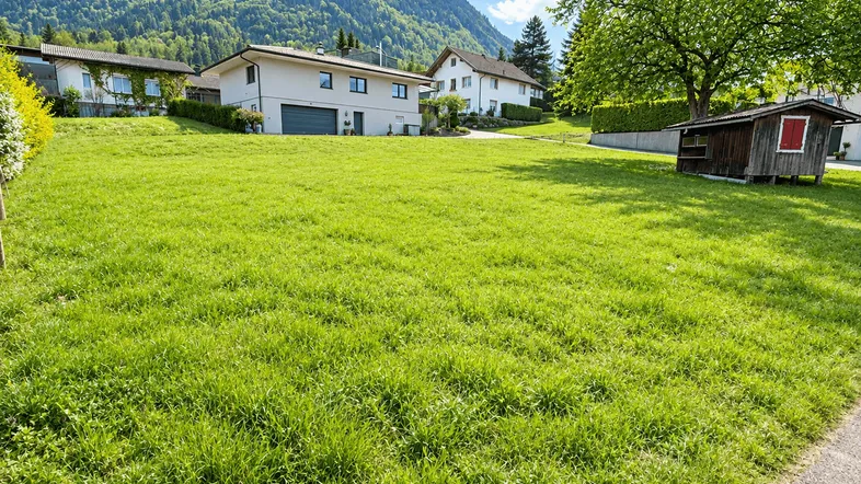 Weitläufige grüne Wiese mit Blick auf moderne Häuser und bewaldete Berge im Hintergrund.