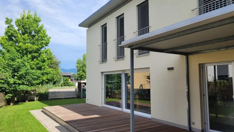 Moderne Hausfassade mit großer Holzterrasse und gepflegtem Garten unter blauem Himmel.