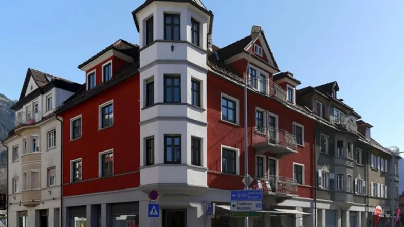 Markantes Eckgebäude mit roter Fassade und spitzem Turm an einer städtischen Kreuzung.