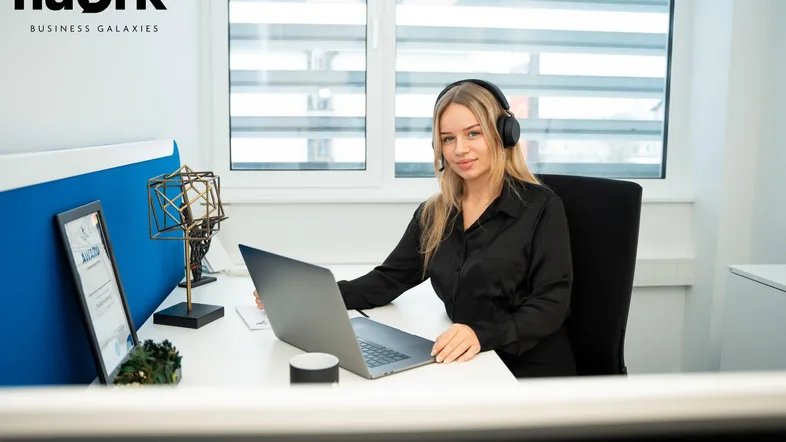 Junge Frau mit Kopfhörern arbeitet an einem Laptop an einem Schreibtisch in einem modernen Büro mit viel Tageslicht.